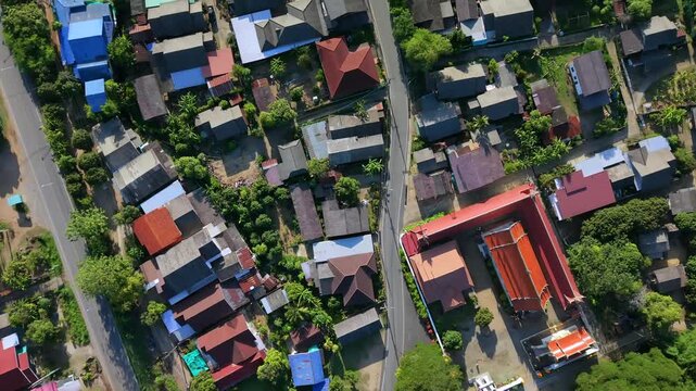 cenital Drone shot flying overhead residential area in a small local town with asphalt curve road houses rooftops home gardens green tropical trees and countryside neighborhood during sunny summer aft