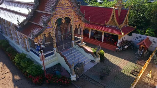 Drone shot rising backwards from the entrance of a traditional Buddhist temple with golden ornaments red roofs naga statues and paved courtyard during a sunny day in Southeast Asia