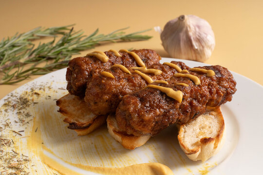 Close-up of Romanian grilled mici sausages with mustard lines, served on toasted bread slices with a background of a garlic bulb and rosemary sprigs.