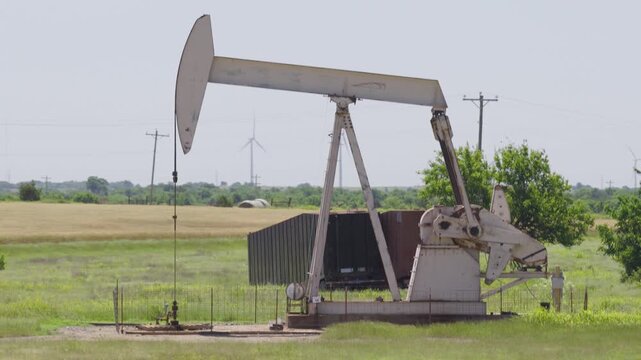 Drone 4K of a wheat field with two oil pump jacks and wind turbines in the background.