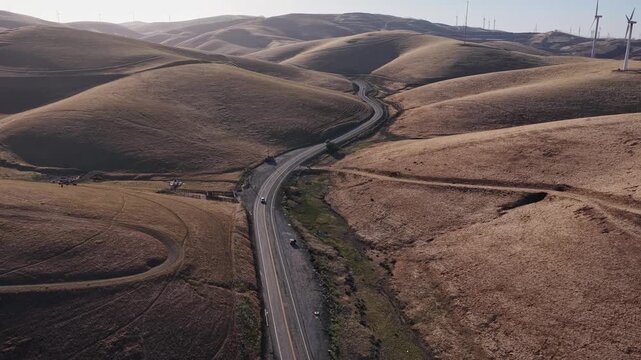 Scenic aerial view of the Altamont Pass wind farm, featuring cars driving on a curvy road through golden brown rolling hills in Livermore, California, a symbol of renewable energy production