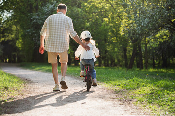 Man helping his child learning to ride bicycle on bike cycling trail in green park, happy family leisure activity, father and daughter together training motor development and activity. © juliasudnitskaya