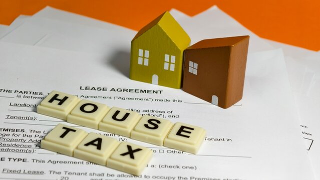 Two small wooden toy houses sit on a lease agreement document, while plastic alphabet letter tiles in the foreground spell out the phrase "HOUSE TAX" against a vibrant orange background.