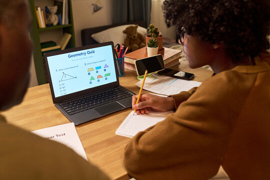 Black teenager studying geometry on laptop, taking notes during online tutoring session at home with parent nearby. Ideal for e learning, homeschool, math education, academic support