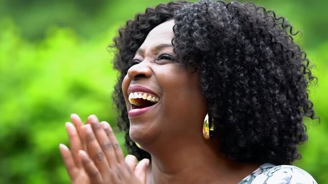 Joyful african american woman, curly hair, gold hoops, black top, laughing, raising fists, then hands interlocked upwards in lush green bokeh, profound celebration