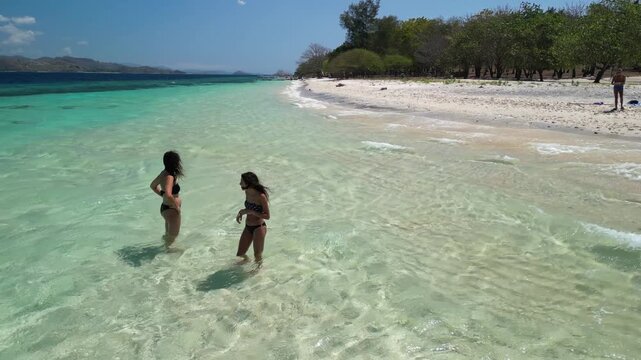 Happy mom and little girl barefoot in transparent sea waves touching sandy shore with gentle waves and beach background