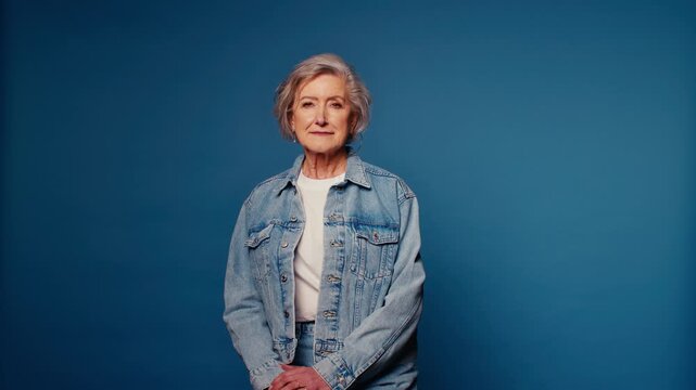 Confident senior woman in denim jacket on blue background. Stylish older female model looking at camera.