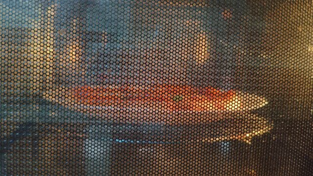 Closeup of pepperoni pizza with circle crust baking inside rotating or spinning hot microwave oven. Making, reheating or preparing simple homemade italian pizza in kitchen concept.
