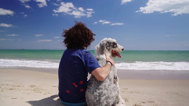 Man and his white and grey shaggy dog seen from behind contemplating the ocean, dog turns towards camera, summer, slow motion.