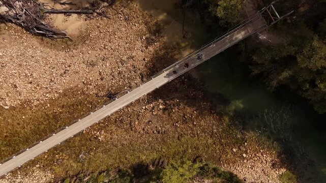 Three hikers walking across a long suspension bridge over turquoise river in lush wilderness of Abel Tasman National Park, New Zealand. Aerial view. Top down.