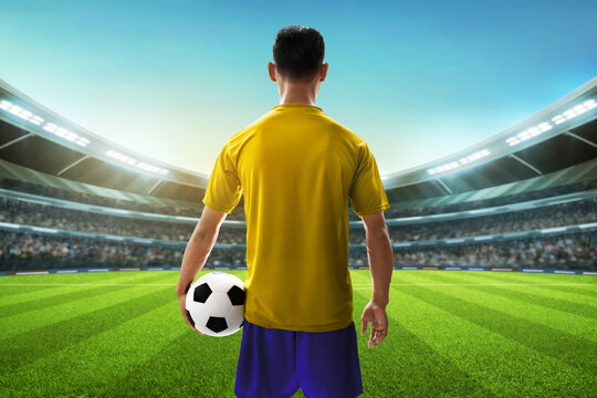 Back view of professional soccer player in yellow and blue uniform holding a ball in the stadium field