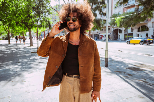 Happy stylish man with afro hairstyle communicating on mobile phone while walking in urban environment.