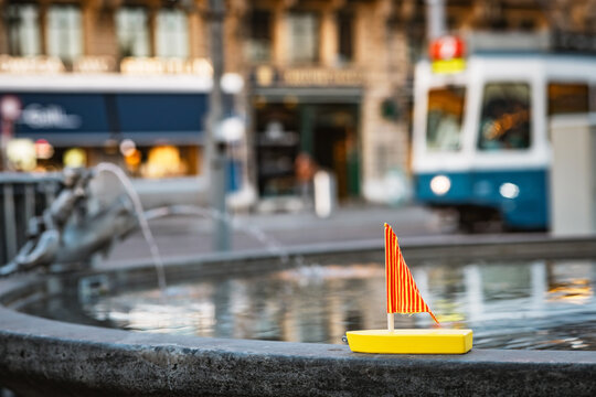 Yellow wooden boat toy placed on a public fountain with drinkable, fresh, mineral spring water tap in Zurich, with a city tram running in front of old buildings and shopping street, Switzerland