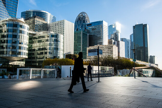 Wide financial district plaza in La Defense, Paris, where a pedestrian crosses between glass towers, sunlight and skyline reflections