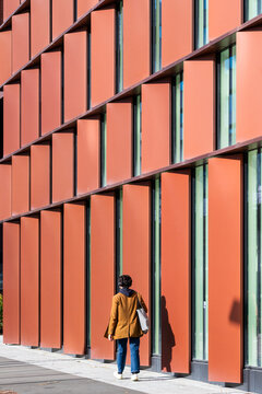Quiet street scene with a person beside a red facade, where vertical panels and shadow give geometric architecture an urban wall presence