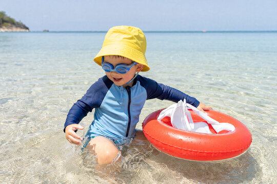 Happy young boy wearing a yellow sun hat and blue goggles plays in the shallow turquoise water of a tropical beach with a red life ring during a sunny summer vacation.