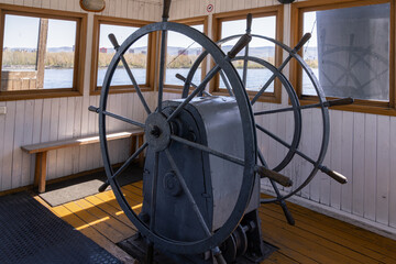 Historic steamship steering wheel inside vintage riverboat wheelhouse with classic navigation mechanism and old maritime interior overlooking river landscape on historic vessel © Vladimir Razgulyaev