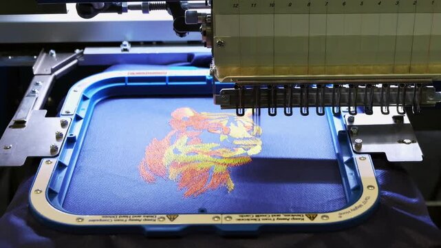 Industrial computerized embroidery machine sewing a colorful lion head design on blue fabric.