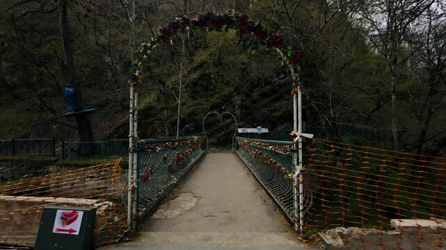 Pedestrian bridge adorned with love locks and an arch decorated with flowers and a heart shape. Bridge leads across a river through a forested area with bare trees and green foliage.