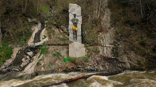 Prometheus statue stands on a rocky cliff overlooking a fast-flowing river. A large log floats in the turbulent water below, with green foliage adorning the hills.