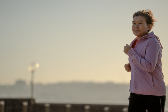 Active senior Asian woman jogging outdoors in misty morning light, healthy lifestyle concept