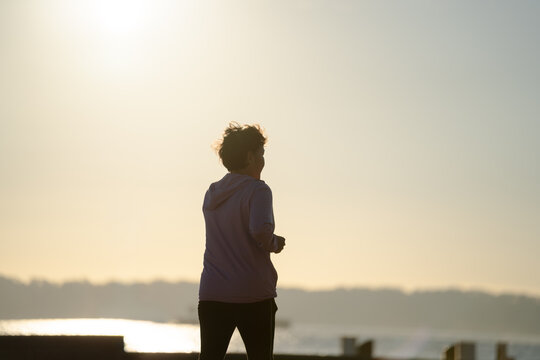 Senior woman jogging along waterfront promenade at sunrise with sun flare, active lifestyle concept