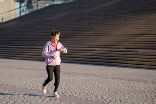 Senior Asian woman checking smartwatch while jogging outdoors, active healthy lifestyle concept