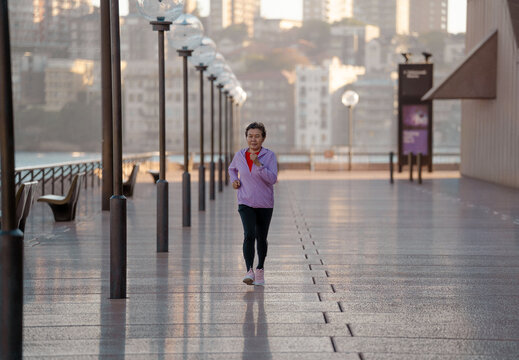 Active senior Asian woman jogging along waterfront promenade at sunrise, healthy lifestyle concept