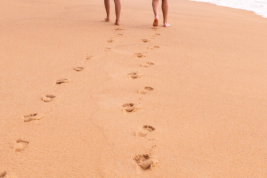 Two pairs of bare feet walk away on a sandy beach, leaving clear footprints in the warm, golden sand.