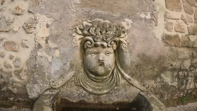 Slow push-in on a weathered stone mascaron relief carved into an ancient Roman wall in Tivoli.