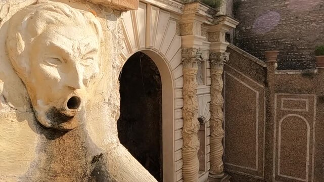 Pan right reveals a weathered lion head stone fountain against ornate baroque columns and ancient walls in Tivoli near Rome.
