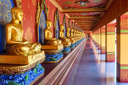 Golden Buddhas lined up in a row inside a Thai temple with intricate red and gold decorations and colorful blue bases, creating a serene and spiritual atmosphere.