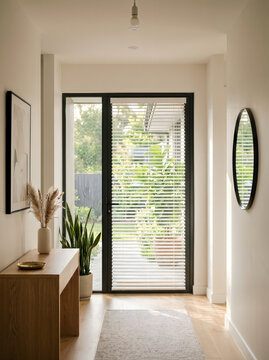 Modern minimalist hallway interior with wooden console table, decorative pampas grass, snake plant, round black mirror, and glass door with blinds leading to a sunny garden