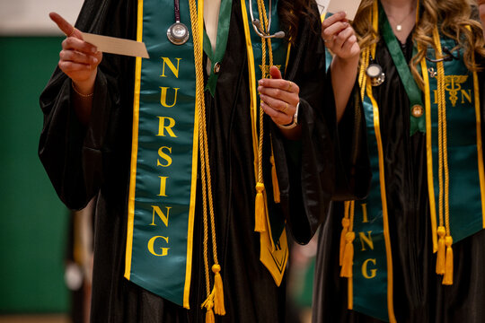 Two nursing graduates in black gowns hold their diplomas while wearing green "Nursing" stoles and stethoscopes at commencement