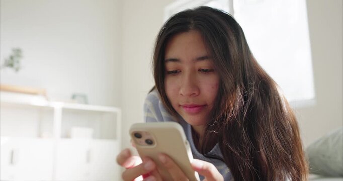 Asian happy young woman on bed using smartphone in soft morning window light at home