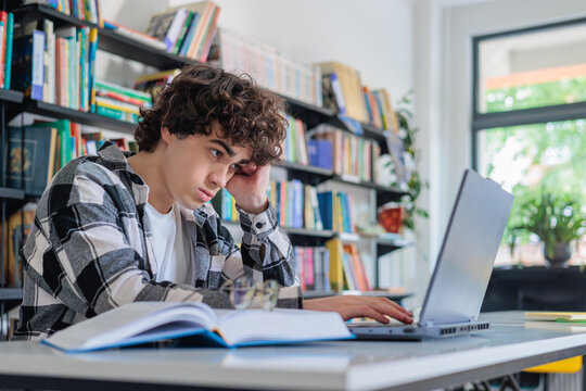 Teenage boy looking bored while studying on laptop in library. Lack of motivation, concentration problems, learning difficulties and student disengagement concept