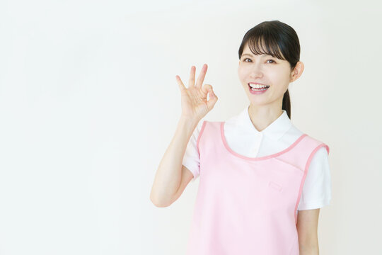 白背景のピンクのエプロンをつけた指で丸のポーズをする若い日本人女性介護士のポートレート