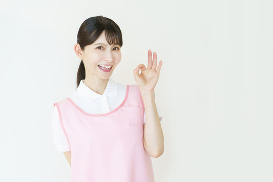 白背景のピンクのエプロンをつけた指で丸のポーズをする若い日本人女性介護士のポートレート
