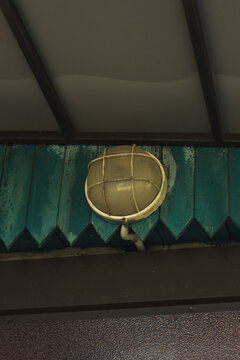 Vintage industrial round wall lamp with protective metal cage mounted on weathered green wooden planks under a ceiling.Old outdoor bulk head light fixture on a peeling turquoise wooden wall