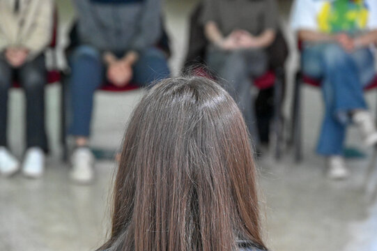A girl sits in front of a group of people. A woman sits opposite other women and men.