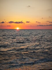 Fototapeta premium Vertical view of sun setting below the ocean horizon. Orange dusk sky with dark clouds and foamy waves