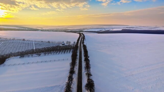 Aerial drone footage of empty road in winter landscape at sunrise, snow covered fields and trees.