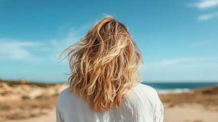 Obraz premium A young woman gazes at the blue ocean, hair flowing in the breeze, capturing a moment of peace and connection with nature in a serene beach setting.