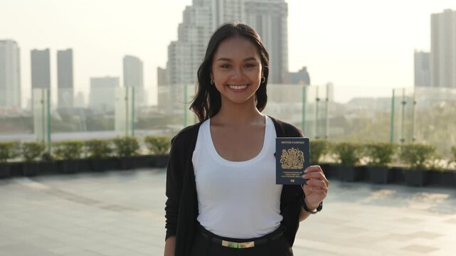 Portrait of cheerful Asian female shows new British passport and stands on city square. Confident businesswoman demonstrates citizenship document and smiles in modern megapolis