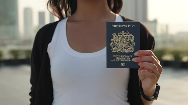 Woman shows passport of British citizen and stands against skyscrapers silhouettes closeup. Lady proud to display personal identification document in summer urban environment
