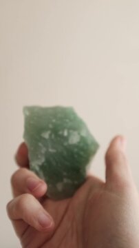 Close-up of a person's hand holding and turning a raw, unpolished green aventurine quartz crystal gemstone against a neutral studio background, vertical orientation.