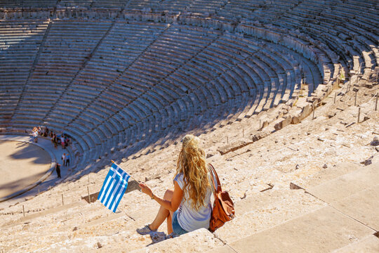 Woman tourist with greek flag sitting on stone steps of ancient theater of Epidaurus, Tour tourism in greece