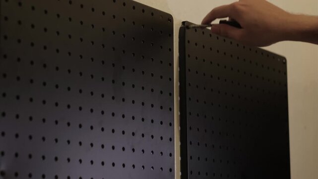 Medium static shot of hands using a screwdriver to install black pegboard panels onto an interior wall
