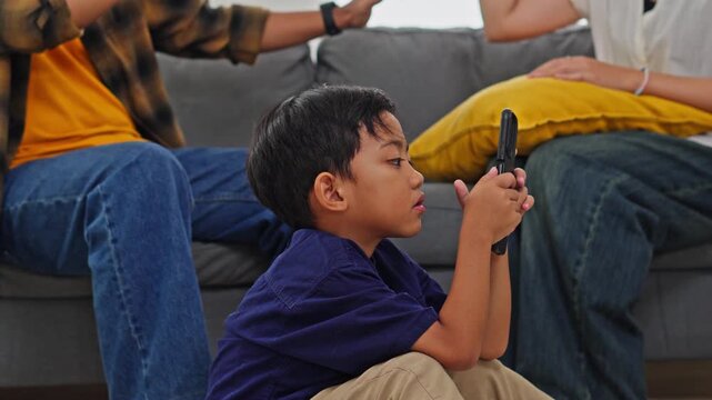 Focused little boy using mobile phone while parents at odds on sofa in background