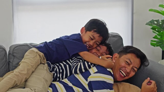 Happy Asian father playing with two young sons while laughing and bonding together on sofa